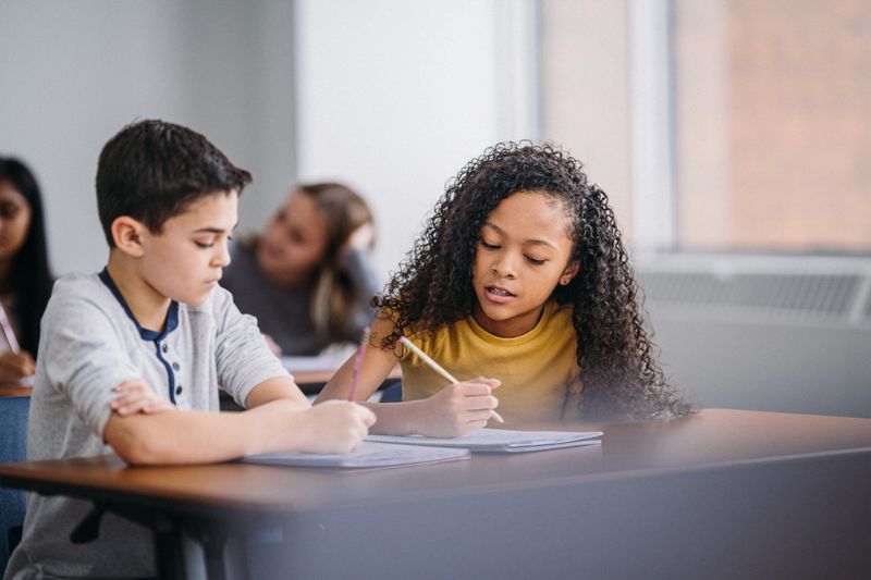 A multiracial middle school boy and girl sit at a classroom table while writing in open notebooks, solving schoolwork problems together.
