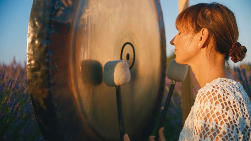 Mature woman engaging in a tranquil sound therapy with a gong, surrounded by a blooming lavender field during a calming sunset.