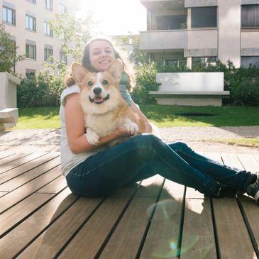 Woman sitting on wooden deck hugging a happy corgi dog in bright sunlight.