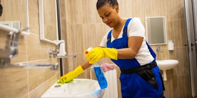 Woman cleaning a bathroom sink wearing yellow gloves and blue overalls.