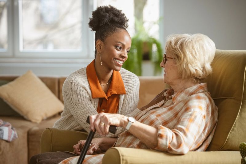 Smiling female home caregiver supporting senior woman in her house, talking with her. Senior lady holding walking cane.