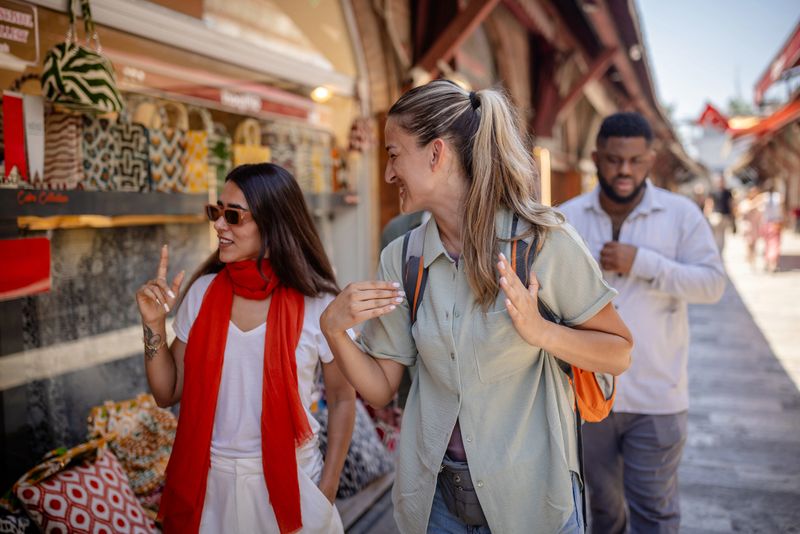 Tourists engaging in an animated discussion and gesturing while exploring outdoor market stalls filled with colorful bags and other unique local products, under the warm sun.