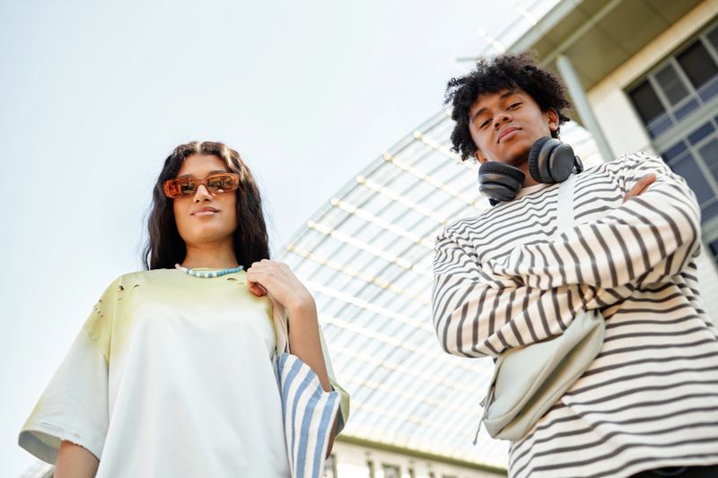 Low angle shot of teenage multiethnic boy and girl standing outside in city looking down at camera while posing against bright summer sky, copy space