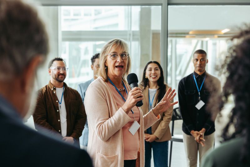A confident female speaker addresses a group of employees during a training session, encouraging engagement and promoting professional growth and team building in a modern office setting.