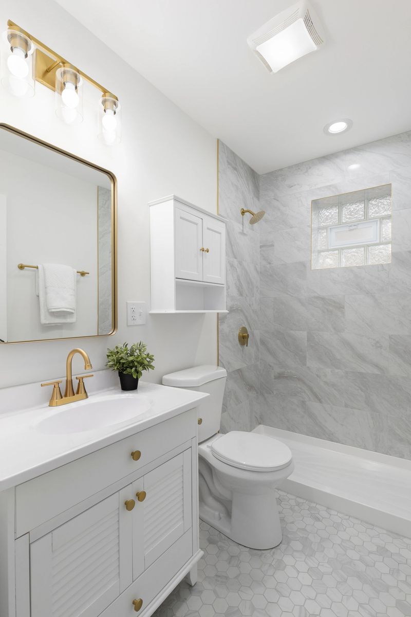 A white bathroom with gold lights, faucet, and light and a shower with marble tiles.