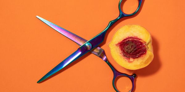 Iridescent scissors next to a halved peach on an orange background.