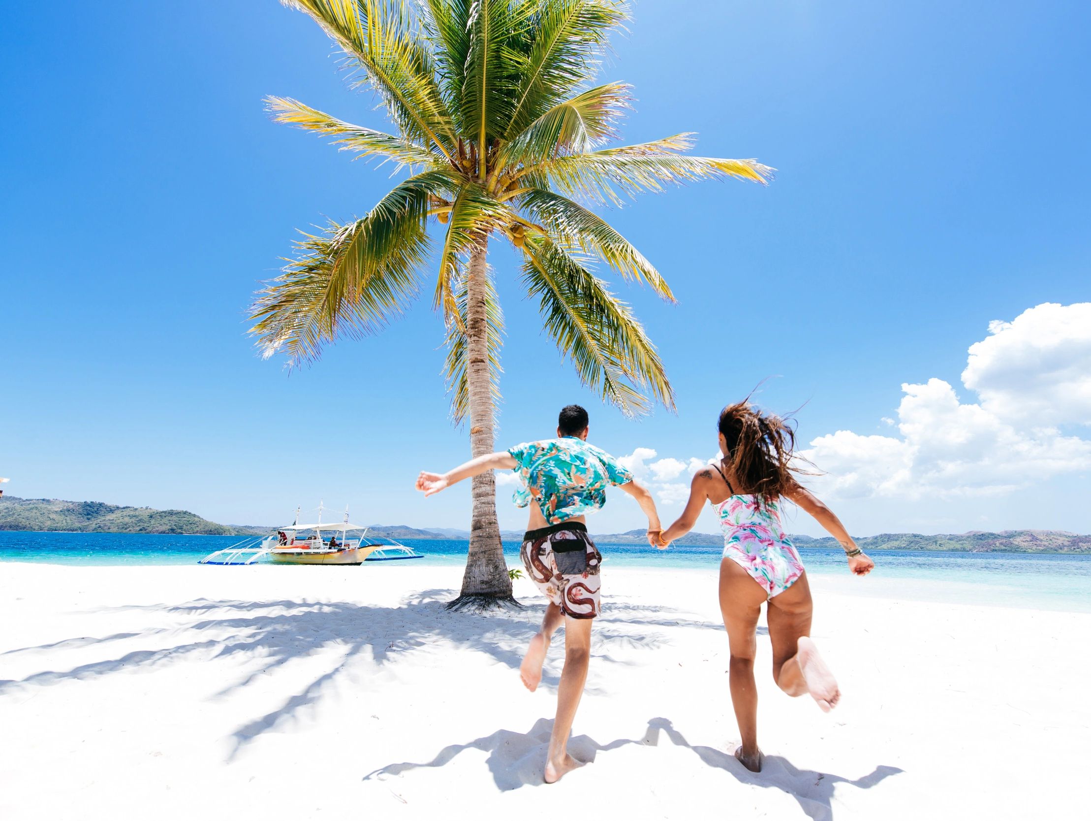 Couple running hand-in-hand on a sunny tropical beach with a palm tree and boat.