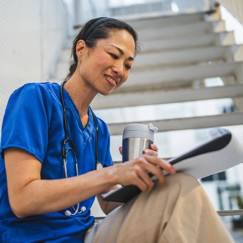A nurse in blue scrubs reviews documents while holding a travel mug.