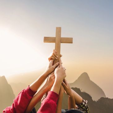 Multiple hands holding a wooden cross against a mountainous sunrise.