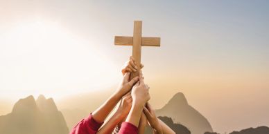 Multiple hands holding a wooden cross against a sunrise mountain backdrop.