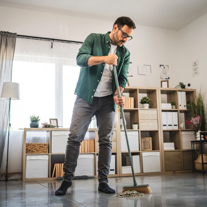 Adult man is sweep the broken vase from floor in a modern, bright living room featuring wooden bookshelves, houseplants, and contemporary furnishings, emphasizing tidy and organized living space