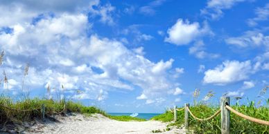 Sandy beach path leading to the ocean under a bright blue sky with fluffy clouds.
