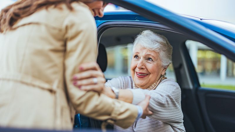 An elderly woman smiles warmly as she is assisted out of a car by a companion. The scene conveys friendship, care, and joy.