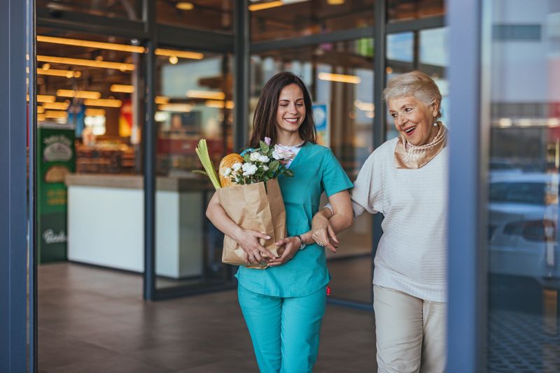 Smiling young caregiver helping elderly woman with grocery shopping in a store. The caregiver carries a paper bag filled with groceries and flowers, highlighting care and companionship.
