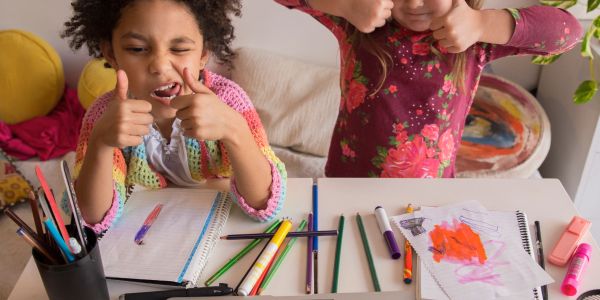 Two kids giving thumbs up while sitting at a table with art supplies.