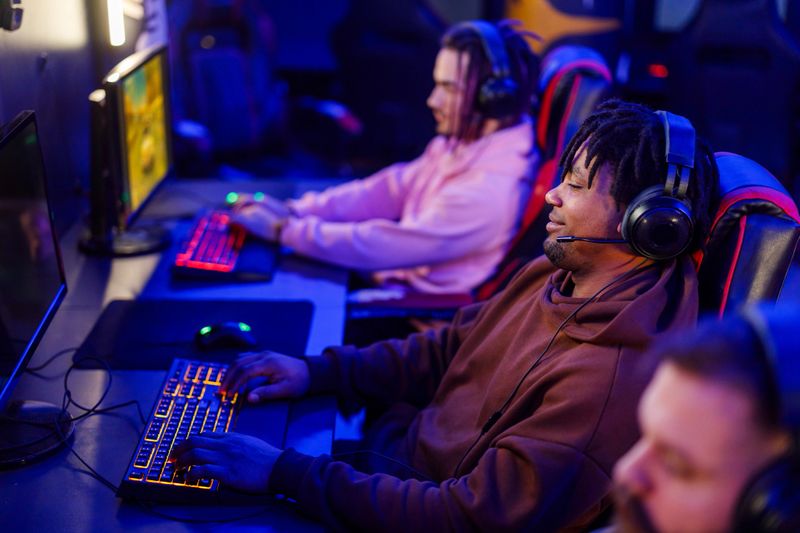 Side view of a professional multicultural gamer sitting in neon lighted gaming lounge with his friends and playing mmo games. A diverse gamer with hands on keyboard is playing online video games.