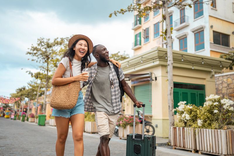 A smiling young Asian woman and a cheerful Black man, dressed in casual travel attire, pull a suitcase and explore a colorful city street together.