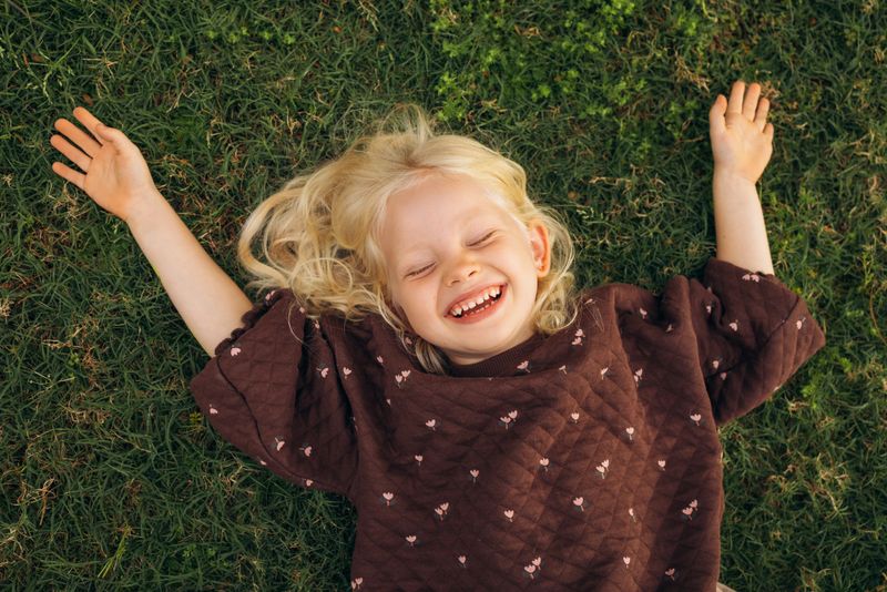 A heartwarming image of a young blonde girl lying on the grass, arms spread wide with delight. She wears a brown sweater adorned with delicate patterns. Her face is animated with a wide, joyous laugh, eyes closed in pure enjoyment. Her tousled blonde hair fans out around her head, blending harmoniously with the green grass. This photograph captures the essence of carefree childhood, showcasing a moment of genuine happiness and playfulness in a serene outdoor setting.
