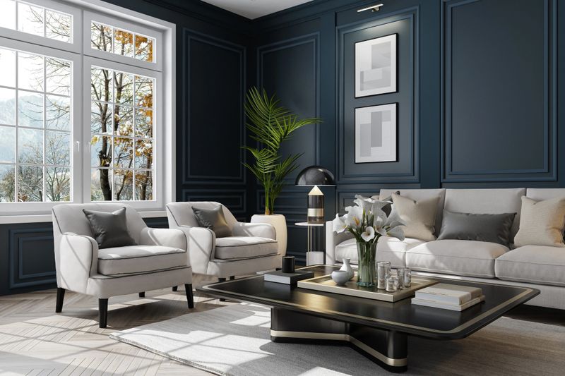 Experience the charm of modern neo-classical Parisian interior design with this luxurious living room setup. The space features classic wall moulding dark royal blue plaster design and large classic windows that flood the room with natural light. The sophisticated white and beige color palette is complemented by plush pillows and sleek modern furniture. A stylish living room table with golden accents and a striking vase filled with white flowers add the finishing touches. This image is ideal for showcasing modern furniture, elegant decor, clean design, cozy room ideas, and renovation projects with a modern classic style.