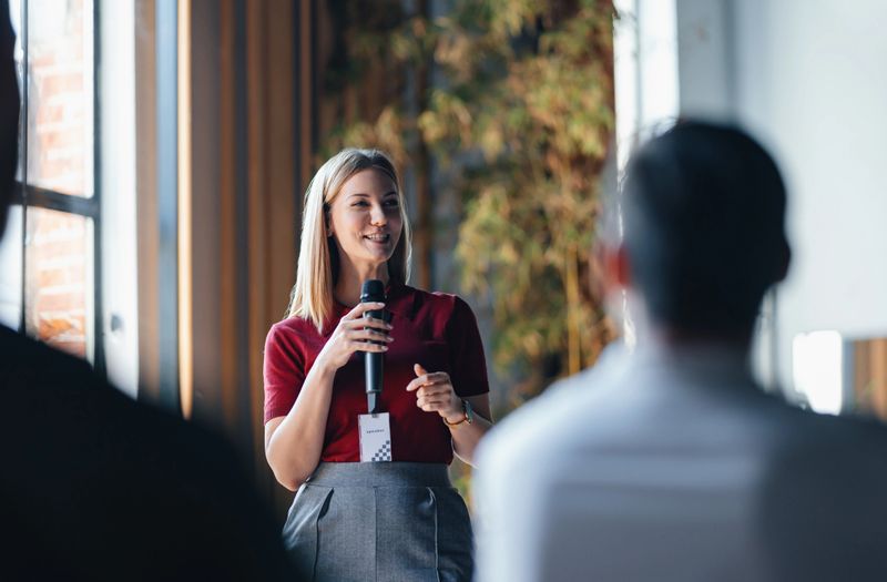 A confident woman delivers a speech to an attentive audience at a professional networking event. The setting suggests collaboration and knowledge sharing among business professionals.