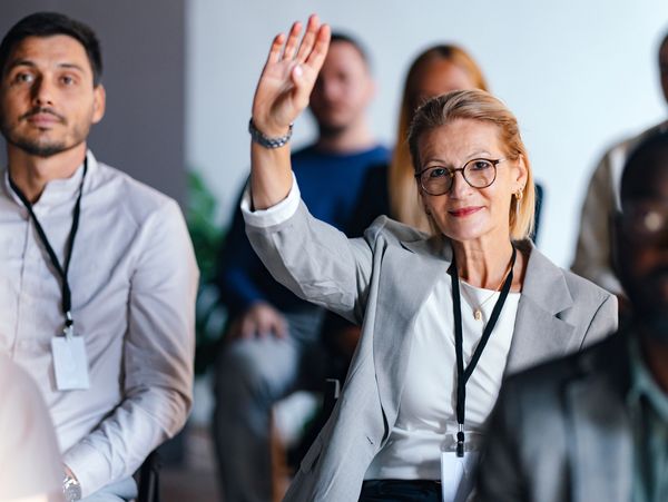 A woman in a gray blazer raises her hand in a meeting full of attentive people.
