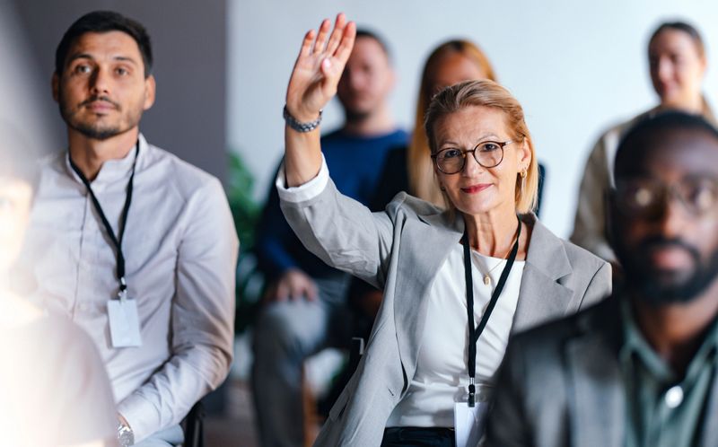 Focused businesswoman raises hand during a professional event, indicating engagement and participation. Diverse group of people in a coworking setting, highlighting teamwork and collaboration.