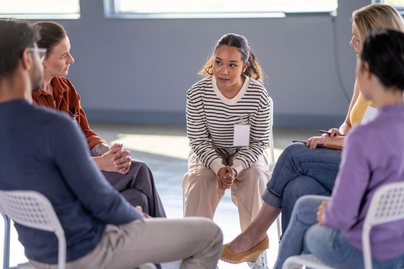 A small group of mature adults sit together in a circle as they meet for a therapy session.  A young female is sharing her struggles with the group as they each listen attentively and show support.