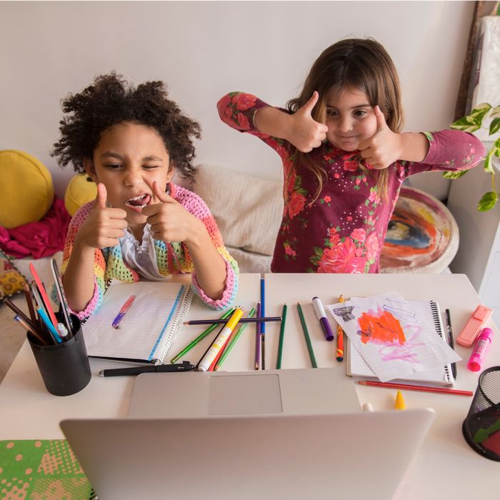 Two young girls give thumbs up while sitting at a desk with art supplies and a laptop.