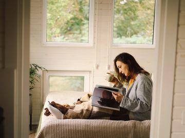 Woman in cozy outfit reading a book on a bed by large windows.