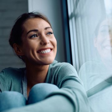 Woman smiling softly while gazing out a bright window, radiating peace, hope, and contentment