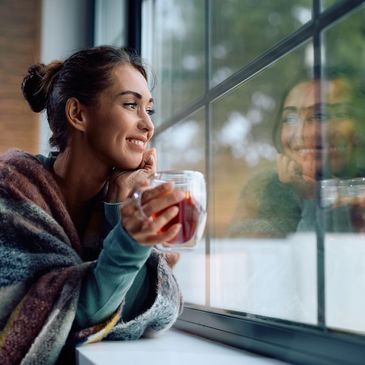 A woman wrapped in a cozy blanket enjoys a warm drink by the window, smiling peacefully.