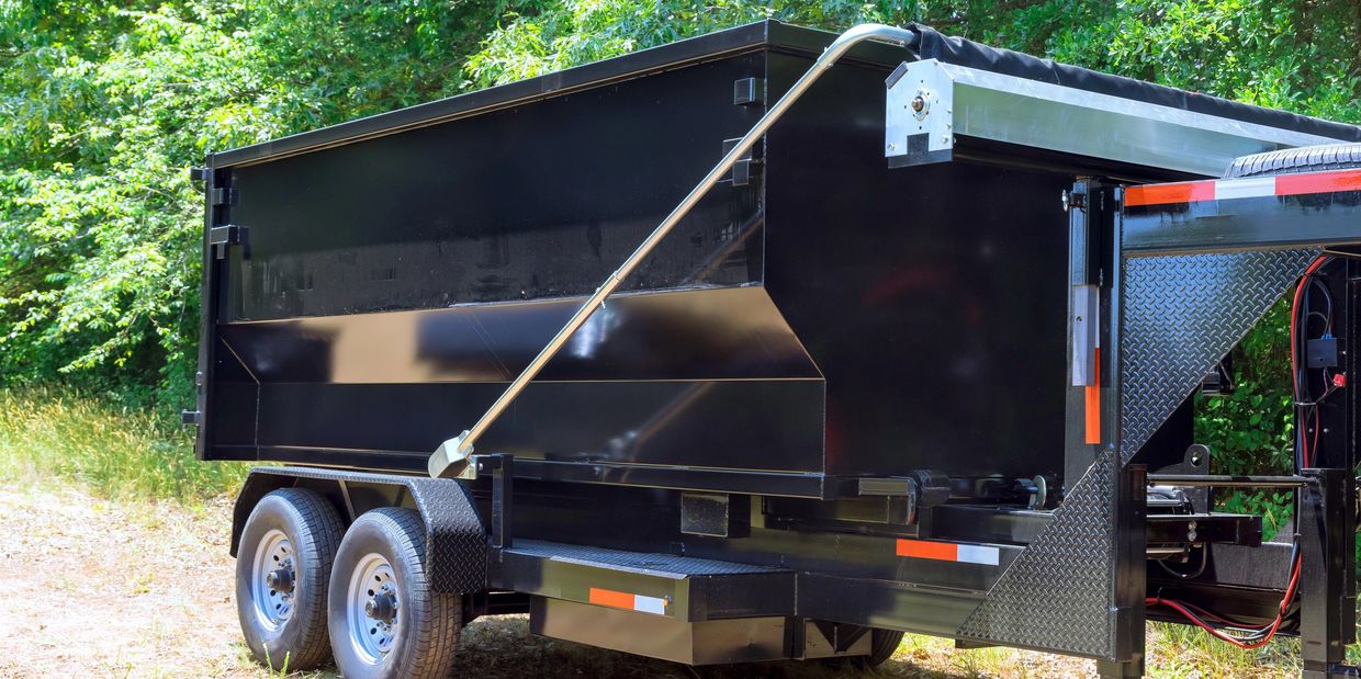 A shiny black dump trailer parked on a dirt path with green trees in the background.