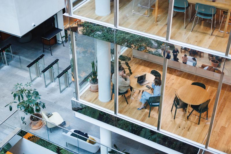 A group of diverse executives engage in a conversation at a stylish café inside a contemporary office, surrounded by glass walls, lush plants, and casual seating arrangements.