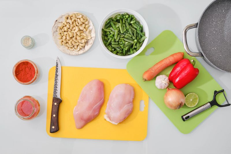 Fresh chicken breasts accompany vibrant vegetables and spices, ready for a flavorful stir-fry. Cashews, green beans, and colorful peppers are arranged beside a pot for cooking.
