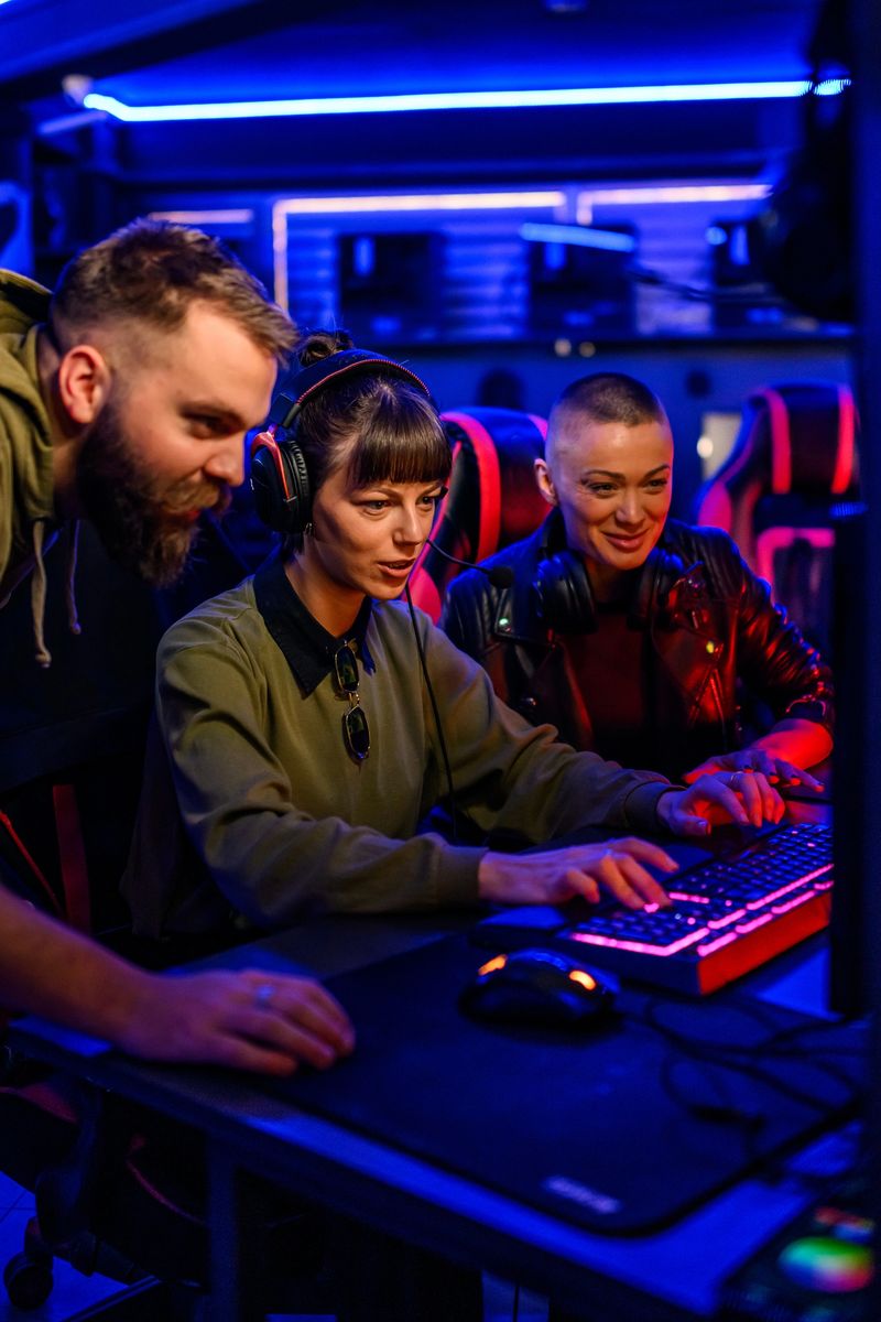 A young gamer girl with headset sitting in gaming lounge and playing a video game while her friends helping her with challenge. A gamer girl is smiling at the monitor and having battle in video game.