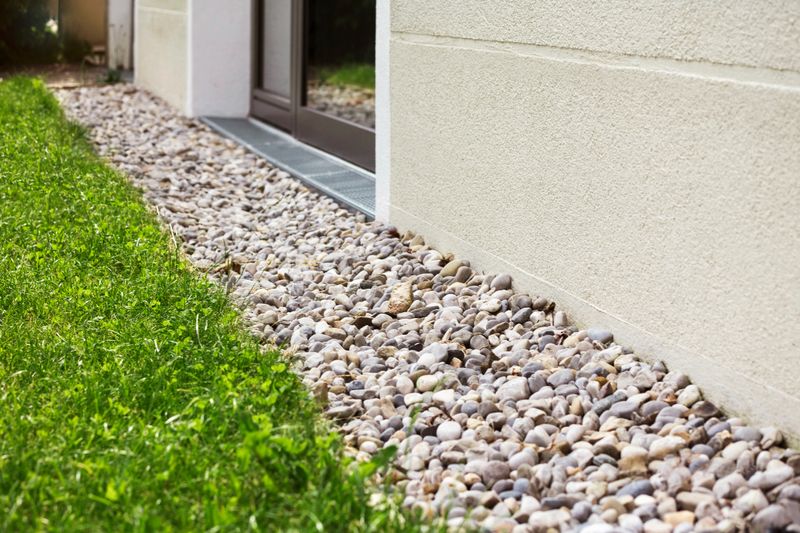 French Drain Gravel Floor along House Wall Outside with Steel Grate by Window Door. Drainage Covering Surface of Crushed Stones Pebbles along Perimeter Modern Building.