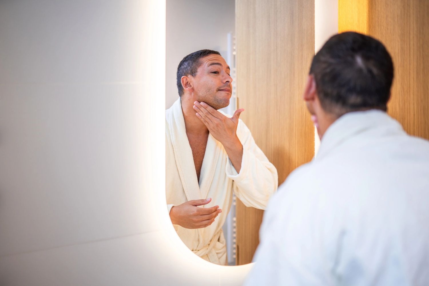 Man in a white bathrobe checking his face in the mirror. Applying body butter after a shower
