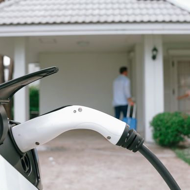 An EV charger handle in EV port with man walking into house in background.