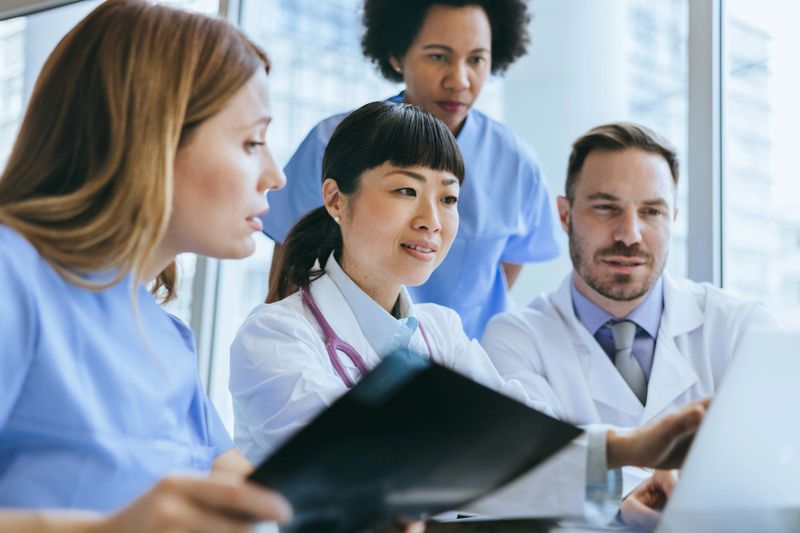 Doctors and nurses are involved in a medical discussion over a laptop in a light-filled hospital meeting room