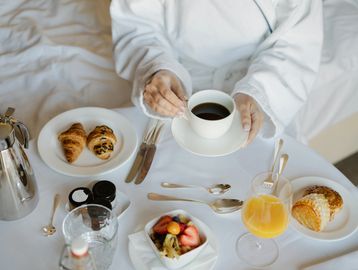 Person in a robe enjoying breakfast in bed with coffee, pastries, fruit, and juice.