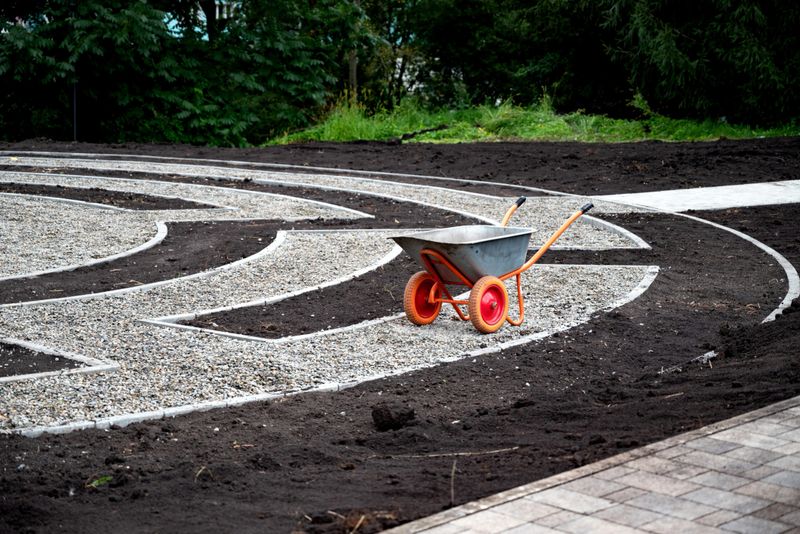 Wheelbarrow on a landscaped garden pathway with gravel and soil sections, lush green background. Concept of gardening and landscaping