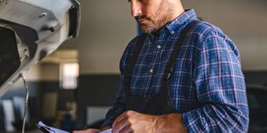 Mechanic in plaid shirt inspecting a car engine with clipboard.