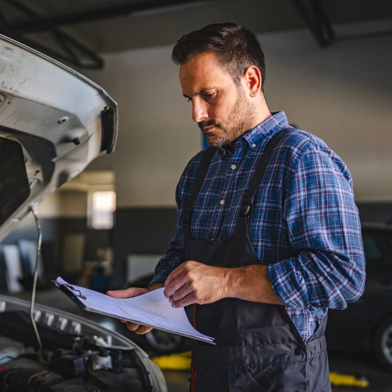 A car mechanic in a car repair garage is examining the open engine compartment and reviewing diagnostics results on a clipboard, signifying diligent vehicle maintenance and service efficiency