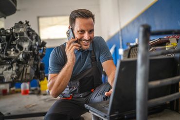Mechanic talking on phone while working on a laptop in a garage.