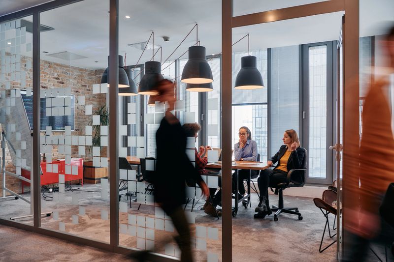 In the dynamic hustle and bustle of a business environment, a group of young business professionals walk down the corridor next to their office, while their colleagues collaborate inside, symbolizing teamwork and productivity.