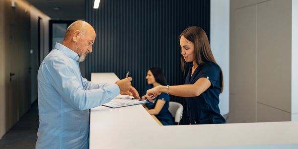 A man signs a document at a reception desk with two female receptionists.