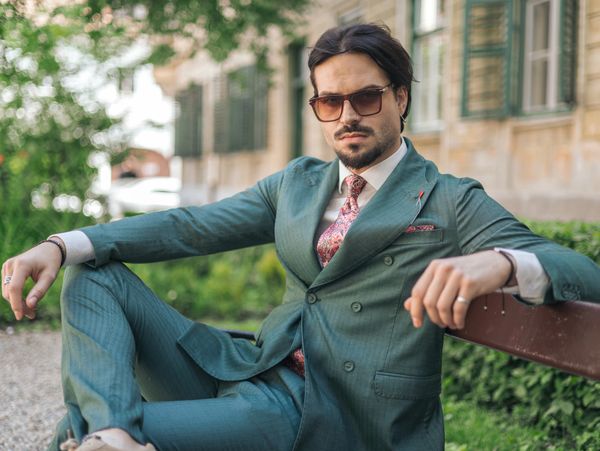Confident man in a teal suit and sunglasses relaxing on a park bench.