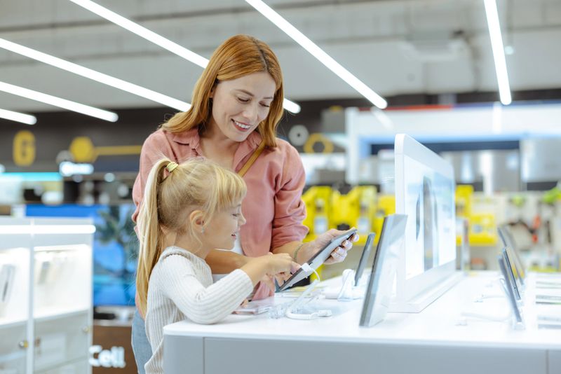 A smiling mother and her daughter interact with a digital tablet at a technology store, highlighting family bonds and modern shopping experiences.
