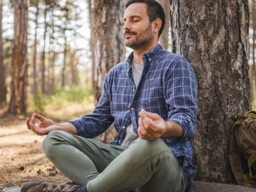 Man meditating outdoors, sitting cross-legged against a tree.
