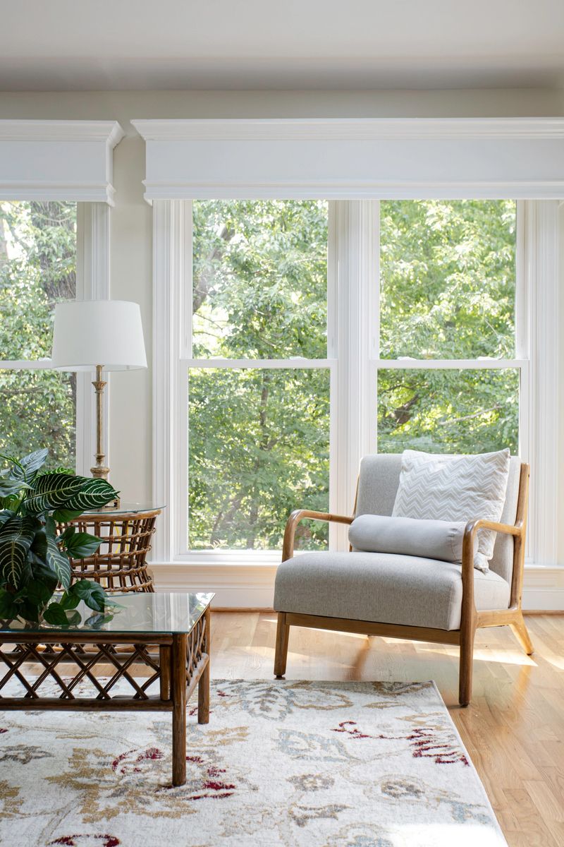 A cozy living room corner with large windows allowing natural light, featuring a grey armchair, an intricate rug, a side table with a decorative plant, creating a peaceful setting.